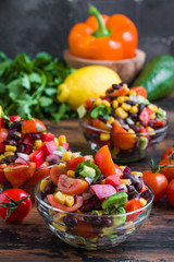 Mexican salad with avocado, tomatoes, beans, corn, red onions and cilantro in glass bowls on a wooden table.