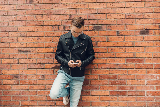 Teenage boy leaning on a brick wall looks at his phone in Sweden