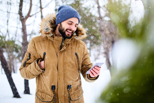Smiling bearded man wears warm winter clothes and using smartphone with fast internet data connection in country side. Handsome man texting with cellphone and using apps in forest. Snowfall in woods.