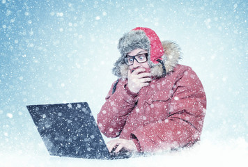 Frozen man in red winter clothes and glasses working on a laptop in the snow. Cold, frost, blizzard, computer