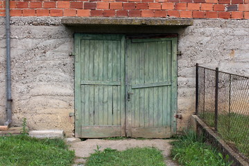 Broken green wooden garage doors with rusted metal hinges and small door handle mounted on concrete wall of red bricks house with uncut grass and metal fence in front