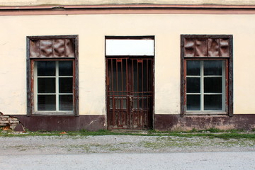 Closed hardware store front with two windows and locked doors with cracked paint mounted on dilapidated broken facade with road and gravel in front
