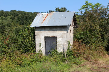 Outdoor garden tool shed made of large concrete building blocks with metal doors and roof surrounded with overgrown plants and forest vegetation on warm sunny day