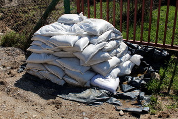 Pile of white sand bags on black nylon cover prepared for flood protection in case of emergency surrounded with gravel and metal fence in background