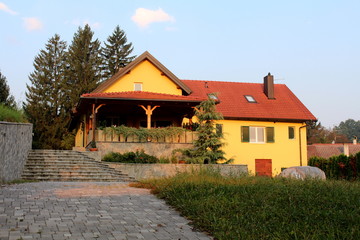 Fototapeta na wymiar Renovated yellow suburban family house with large porch in front, wooden window blinds, new roof tiles, metal chimney and stone tiles driveway surrounded with high uncut grass, flowers and tall trees 