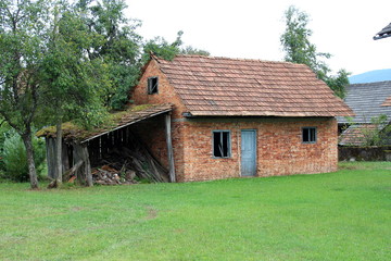 Small abandoned red brick house with broken windows and doors next to old destroyed wooden garage surrounded with freshly cut grass and trees in background