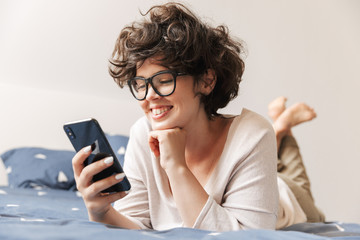 Fototapeta premium Cheerful cute young woman lies indoors on bed chatting by mobile phone.