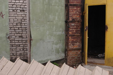 A wall with metal, white brick and red brick parts, metal opened door with wooden planks at a foreground 