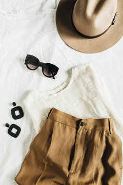 Fashion pastel composition with female clothes and accessories. Beige trousers, blouse, hat, sunglasses, earrings on white blanket. Flat lay, top view.