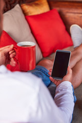 Man sitting on a terrace sofa and drinking coffee/tea while using cellphone.