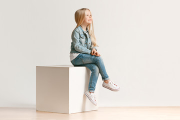 The portrait of cute little kid girl in stylish jeans clothes looking at camera and smiling, sitting against white studio wall. Kids fashion concept - Powered by Adobe