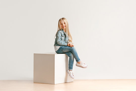 The portrait of cute little kid girl in stylish jeans clothes looking at camera and smiling, sitting against white studio wall. Kids fashion concept