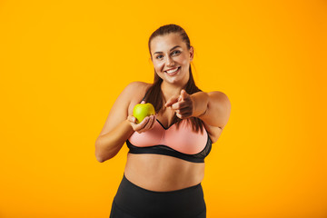 Portrait of healthy chubby woman in sportive bra holding apple, isolated over yellow background