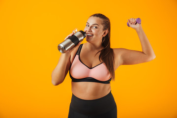 Portrait of young chubby woman in sportive bra drinking water from thermos while lifting dumbbell, isolated over yellow background