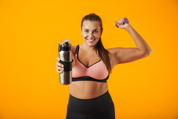 Portrait of european chubby woman in sportive bra holding thermos with water while lifting dumbbell, isolated over yellow background