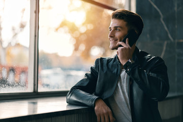 Happy man standing by the window and talking to someone over smart phone