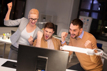 business, success and teamwork concept - happy coworkers with computer celebrating victory at night...