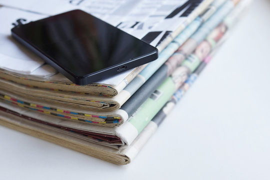 Newspaper and smartphone. Daily papers with news and black mobile phone with touch screen. Modern gadget and folded journal with articles. Electronic device on top of stacked pages, selective focus
