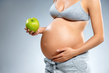 Close up of pregnant woman with green apple and touching her belly on grey background. Pregnancy, maternity, preparation and expectation concept