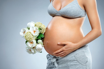 Close up of pregnant woman with bouquet of flowers on grey background. Pregnancy, maternity, preparation and expectation concept