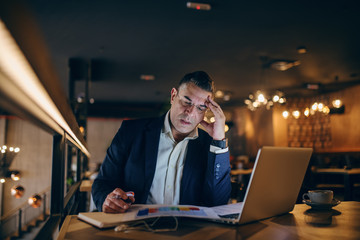 Concerned middle-aged businessman looking at chart and holding pen in hand while sitting in cafe at the evening. On the table coffee, laptop and agenda.