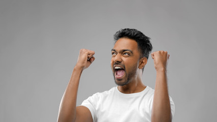 success, emotion and expression concept - happy young indian man celebrating victory over grey...