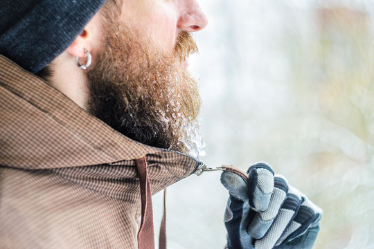 Young man winter portrait in profile. Gloved hand zips a jacket under the chin Beard covered with icicles and frozen droplets. Frost concept