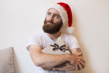 Dreamy man wearing Santa hat and embracing gift box. Bearded guy looking away. Christmas gift concept. Front view.