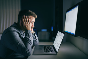 Tired young Caucasian employee holding head in hands and looking at computer monitor while sitting late at night in the office.