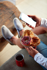 Man sitting on a terrace sofa and drinking coffee/tea, eating donuts while using cellphone.