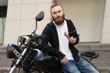 Portrait of confident bearded man standing reclining on bike. Young Caucasian biker holding sunglasses standing at motorcycle outdoors. Biker style concept