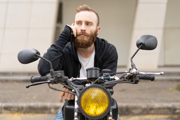 Fototapeta na wymiar Portrait of serious young biker talking on mobile phone outdoors. Caucasian bearded man sitting on motorcycle outdoors. Biker man concept