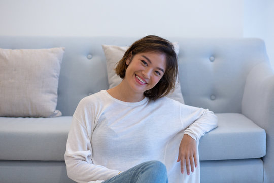 Happy Asian Student Enjoying Weekend At Home. Beautiful Young Woman Leaning On Light Blue Couch With Clasped Hands And Smiling At Camera. Sweet Home Concept