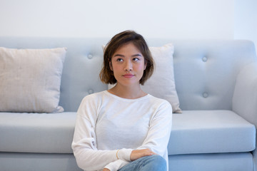 Pensive Asian girl posing at home. Beautiful young woman sitting in front of light blue couch with clasped hands and looking away. Home concept