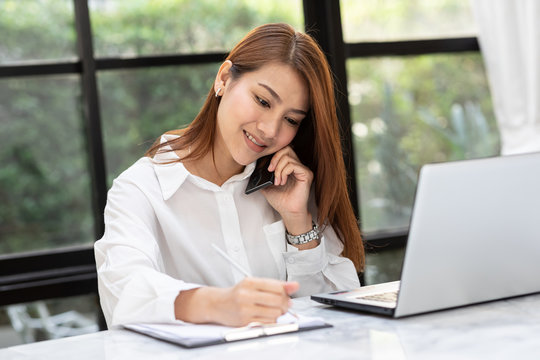 Beautiful Attractive Asian Business woman writing down requirement to book and talking with customer in smartphone,Feeling so happiness and working with service mind,Business and Finance Concept