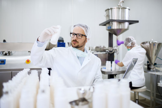 Chemist holding and looking at bottle with liquid soap while doing quality control. On desk bottles and laptop. In background his assistant pouring soap in bottles.