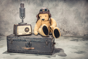 Retro radio, studio microphone, Teddy Bear toy with leather aviator's hat and goggles sitting on old aged classic travel trunk circa 1900s. Broadcasting concept. Vintage style filtered photo