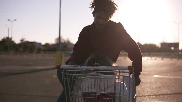 Afroamerican young boy with trendy haircut sitting on a shopping cort on the ouside parking zone and looking to his girlfriend. Caucasian girlfriend sitting inside the cort and talking to her