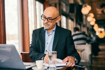 Fototapeta na wymiar Bearded senior businessman looking at laptop and writing tasks in agenda while sitting in cafeteria. On the desk cup of coffee and glass of water.