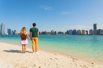 Young couple of tourists holding hands on beach looking at the modern buildings from Abu Dhabi. Honeymoon excursion and summer travel concept with young people travelers relaxing at trip vacation