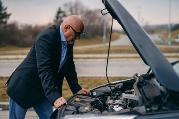 Bearded senior adult man standing in front of opened hood of his car and trying to fix engine.