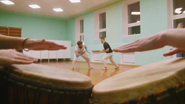 Playing a two musical instruments atabaque on background male fighter performs martial tricks with dance elements in the sport gym