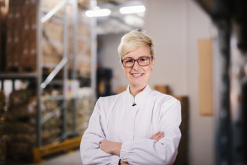 Happy Caucasian female worker posing in warehouse. Arms crossed. In background boxes on shelves.