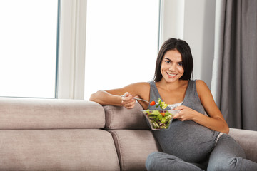 Pregnant woman indoors at home sitting on sofa eat salad.