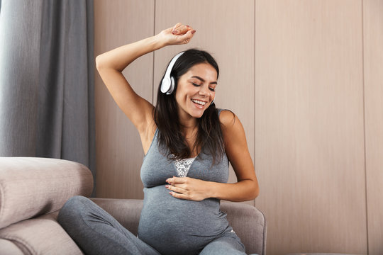 Healthy pregnant woman indoors at home sitting on sofa listening music with headphones.