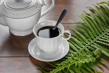 On the table is a cup of black coffee. Nearby is a teapot and a green leaf of a fern.