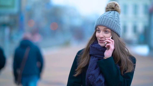 A young girl walking down the street and talking on the phone. Near ride cars. Winter and cold. Beautiful hat.