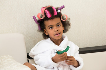 Black girl with curlers with hairpins in her hair. A little girl in a white coat spun pink curlers...