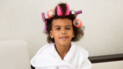 Black girl with curlers with hairpins in her hair. A little girl in a white coat spun pink curlers into her hair.