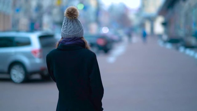 A young girl walking down the street and talking on the phone. Near ride cars. Winter and cold. Beautiful hat.